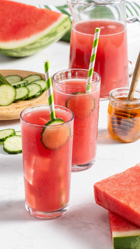 Two watermelon cucumber drinks with pitcher, fresh cucumber and fresh watermelon in the background