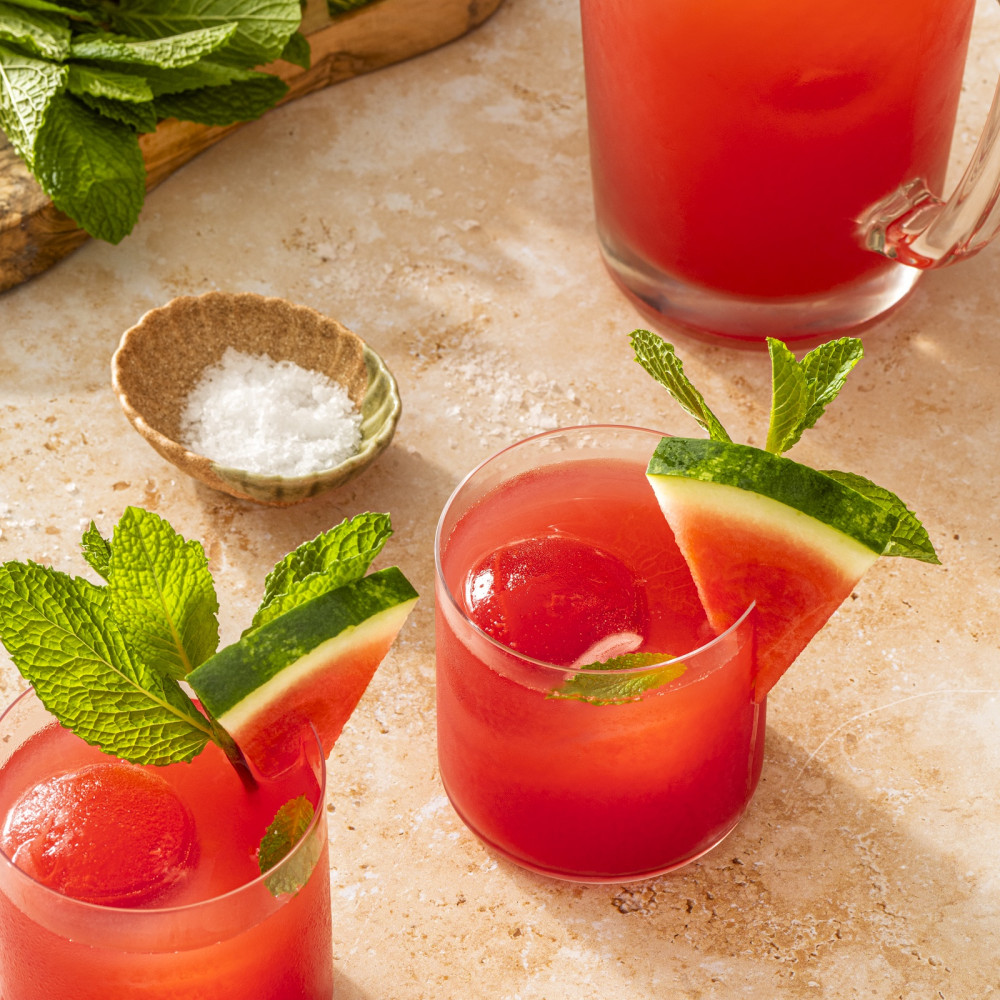 Two cups of watermelon juice with watermelon ice cubes garnished with watermelon slices. Small bowl of salt and pitcher with watermelon juice off to the side.