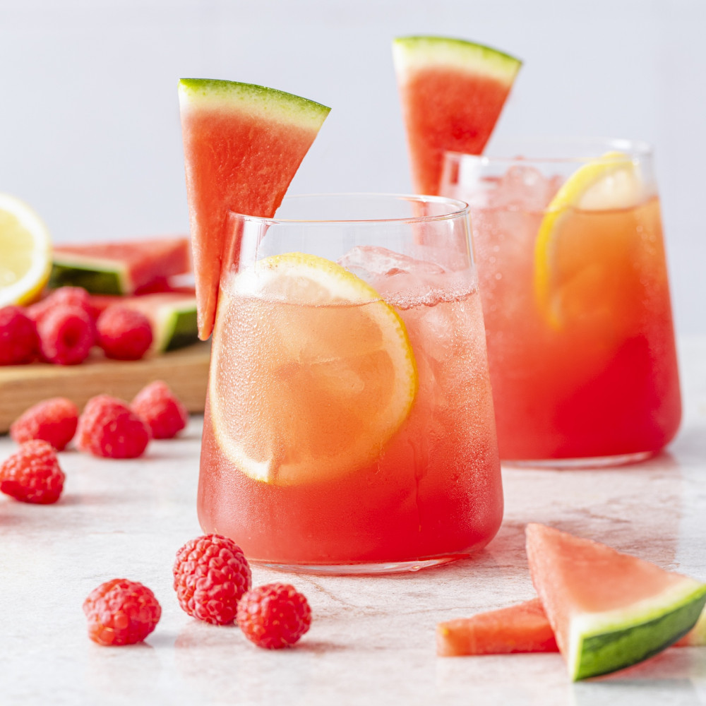 Two cups with watermelon raspberry lemonade with watermelon wedge garnish and scattered raspberries.