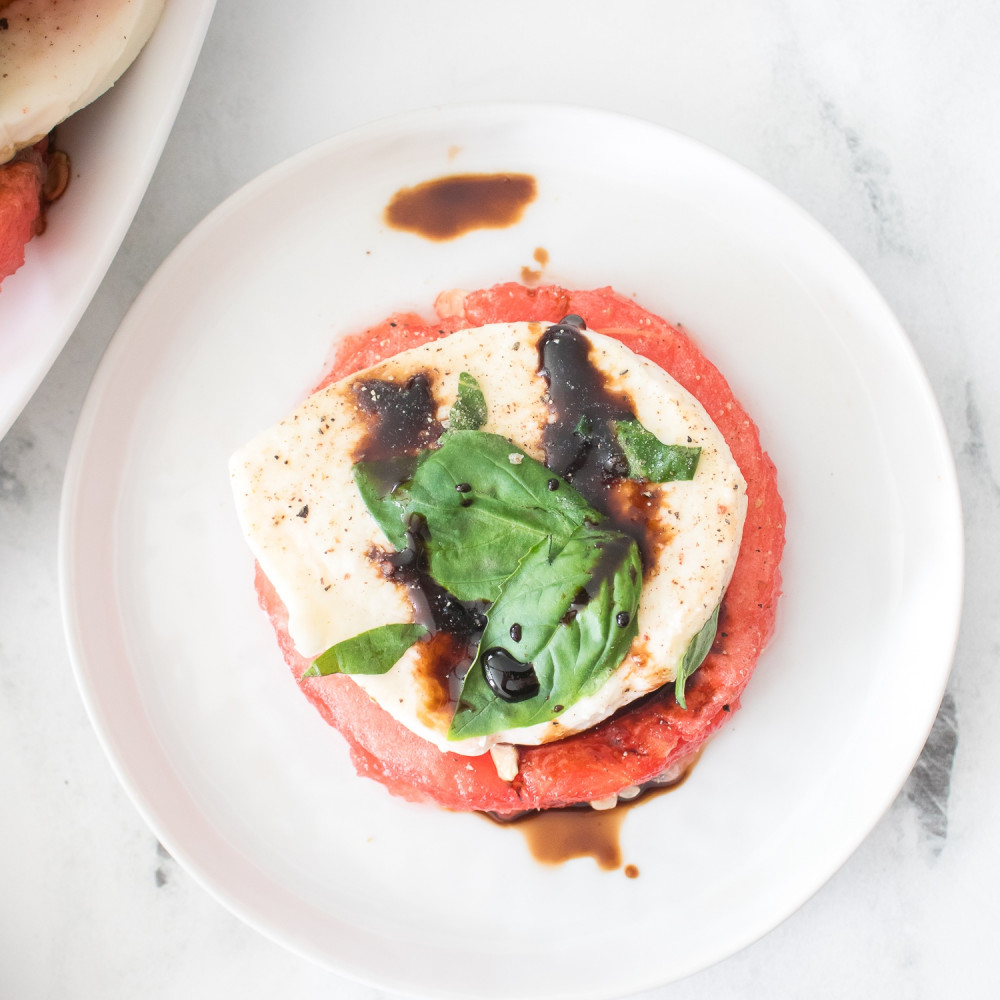 Individual serving of watermelon caprese salad, topped with balsamic and fresh basil, on a white round plate.