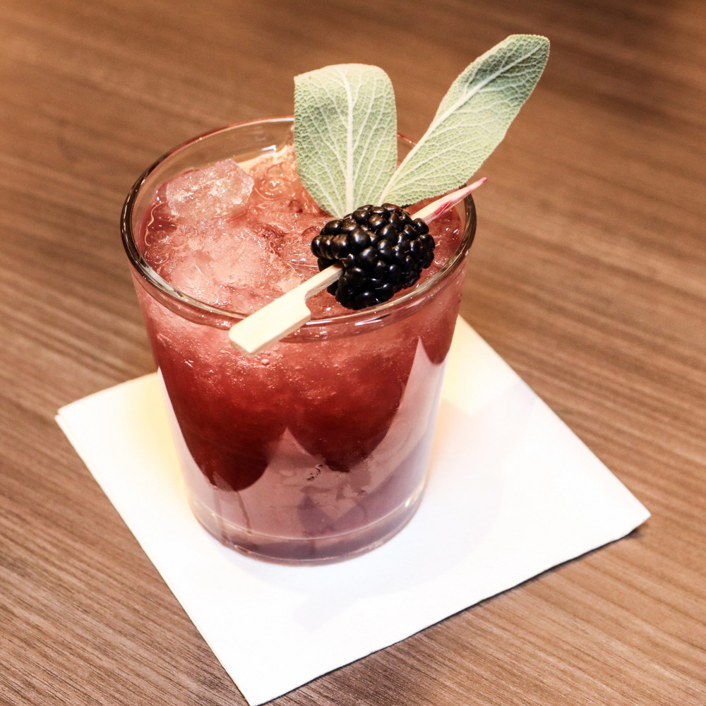 Watermelon and blackberry cocktail in a clear glass with crushed ice on a napkin and topped with toothpick with blackberry and sage leaf.