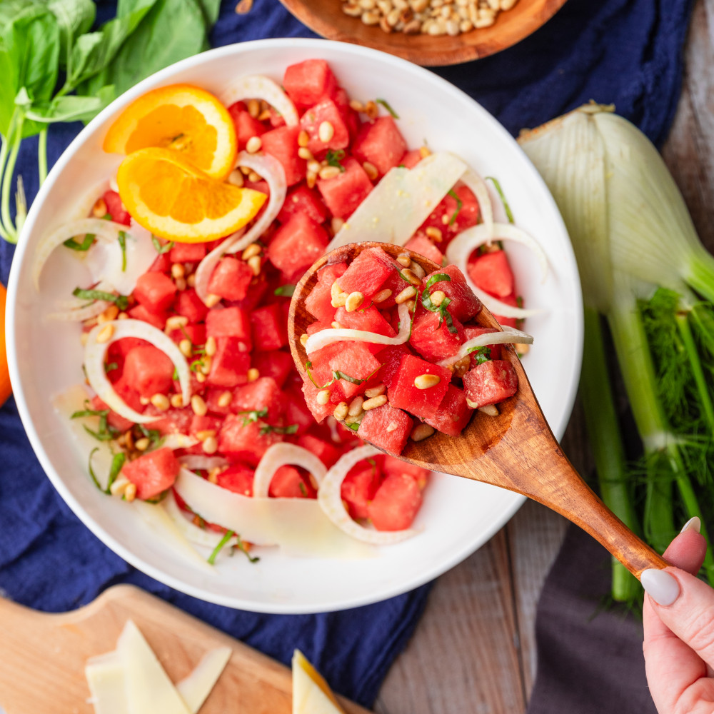 Hand holding a spoon into a bowl containing watermelon fennel salad, surrounded by ingredients including parmesan, fennel, pine nuts, basil and orange juice.