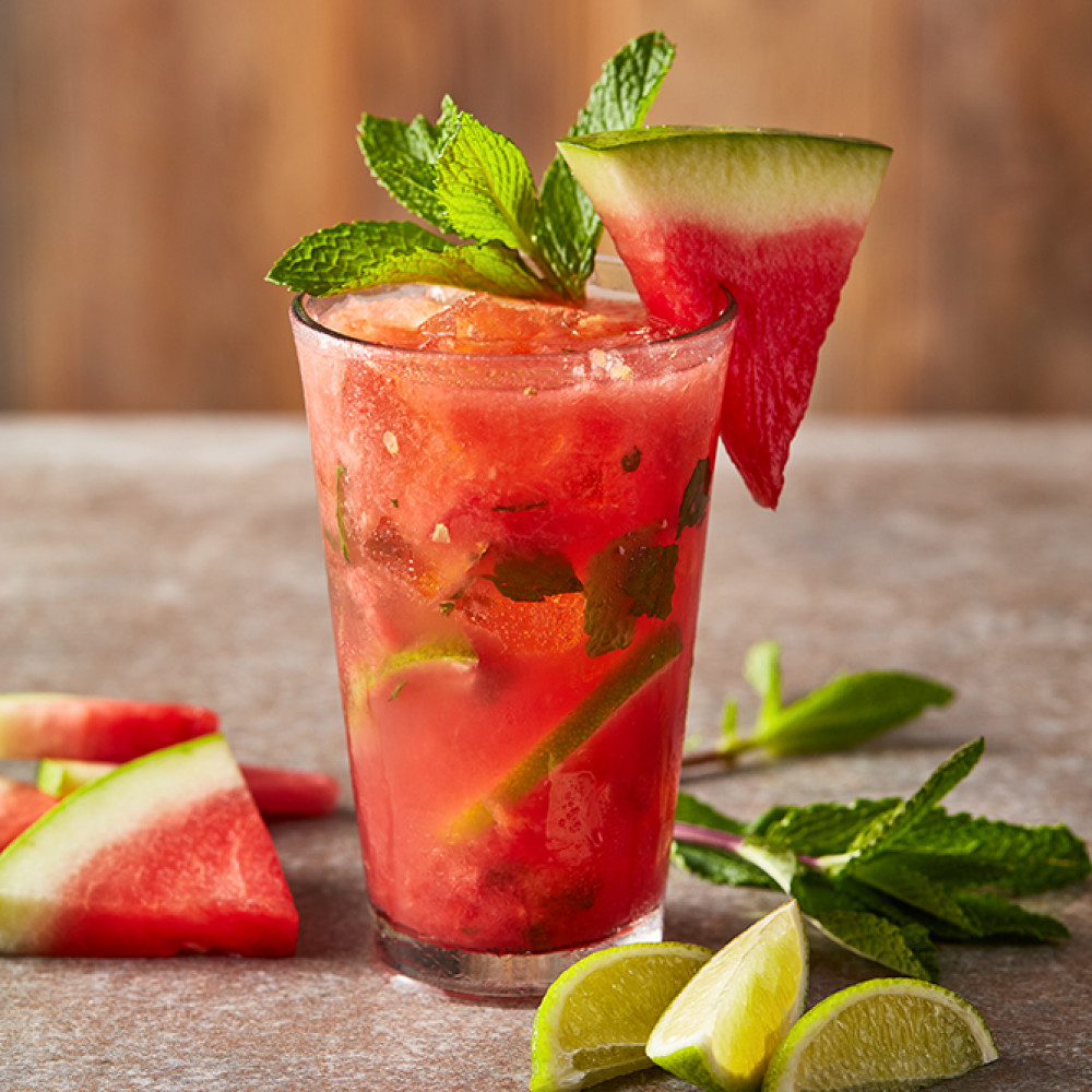 Smokey Mezcal Watermelon Mojito beverage in a tall glass with watermelon wedge, fresh mint and lime wedges.